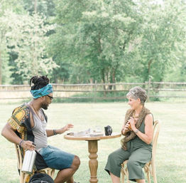 Smiling man and woman sit at a small round table on a sunny grassy lawn, sharing coffee and cards with trees and a rustic wooden fence in the background.