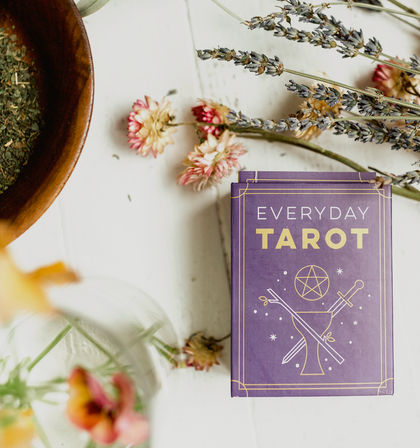 Boho flat-lay of a purple tarot deck box on a white wooden tabletop with dried lavender sprigs, pink strawflowers, a glass vase and a wooden bowl of dried herbs
