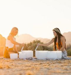 Two women seated on a blanket in a desert at sunset playing white crystal singing bowls in an outdoor sound-bath meditation