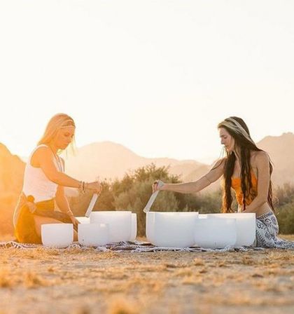 Two women playing white crystal singing bowls on a blanket in a desert at sunset — outdoor sound bath and meditation with mountains and scrub brush in the background.