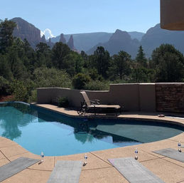 Backyard pool with chaise lounge and yoga mats on the deck, water bottles nearby, framed by pine trees and red‑rock mountains under a clear blue sky.