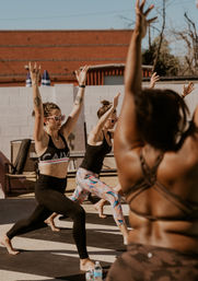 Sunny outdoor urban yoga class with women in athletic wear and heart-shaped sunglasses practicing warrior pose on mats in a courtyard.