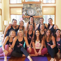 Smiling group of women in matching flamingo tank tops seated on yoga mats in a sunlit cabin living room with wooden floors, large windows and a stone fireplace — cozy indoor yoga retreat scene.
