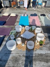 Sunlit rooftop sound-bath and meditation setup with crystal singing bowls arranged on a woven mat around a fluffy cushion, surrounded by flowers and small plants, with colorful yoga mats and pillows laid out for a group session.