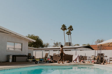 Sunny backyard poolside yoga class with an instructor walking past participants lying on mats on the pool deck, tall palm trees and mid-century houses in the background.