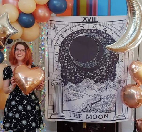 Indoor tarot-themed celebration: large "The Moon" tarot tapestry backdrop with crescent moon and star foil balloons, peach and navy balloon garland, and a guest in a celestial dress holding a rose-gold heart balloon.