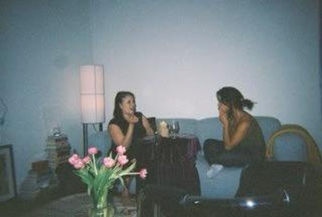 Two friends chatting on a blue sofa in a cozy living room, a small table with wine glasses and a lit candle between them, pink tulips in a vase in the foreground and a soft floor lamp glow.