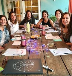 Group of smiling women at a cozy indoor craft night around a long wooden table with wine glasses, rose metal tumblers, paper wheel diagrams, and glowing string lights.