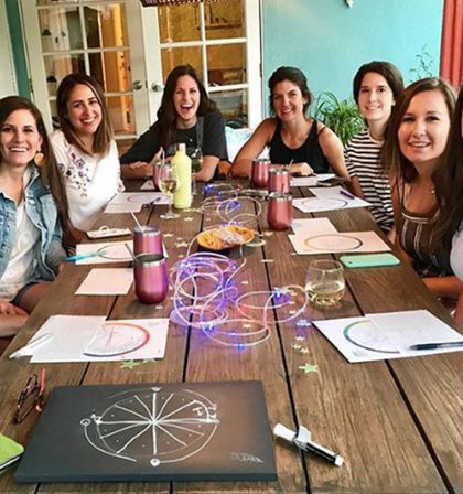 Group of smiling women at a cozy indoor craft night around a long wooden table with wine glasses, rose metal tumblers, paper wheel diagrams, and glowing string lights.