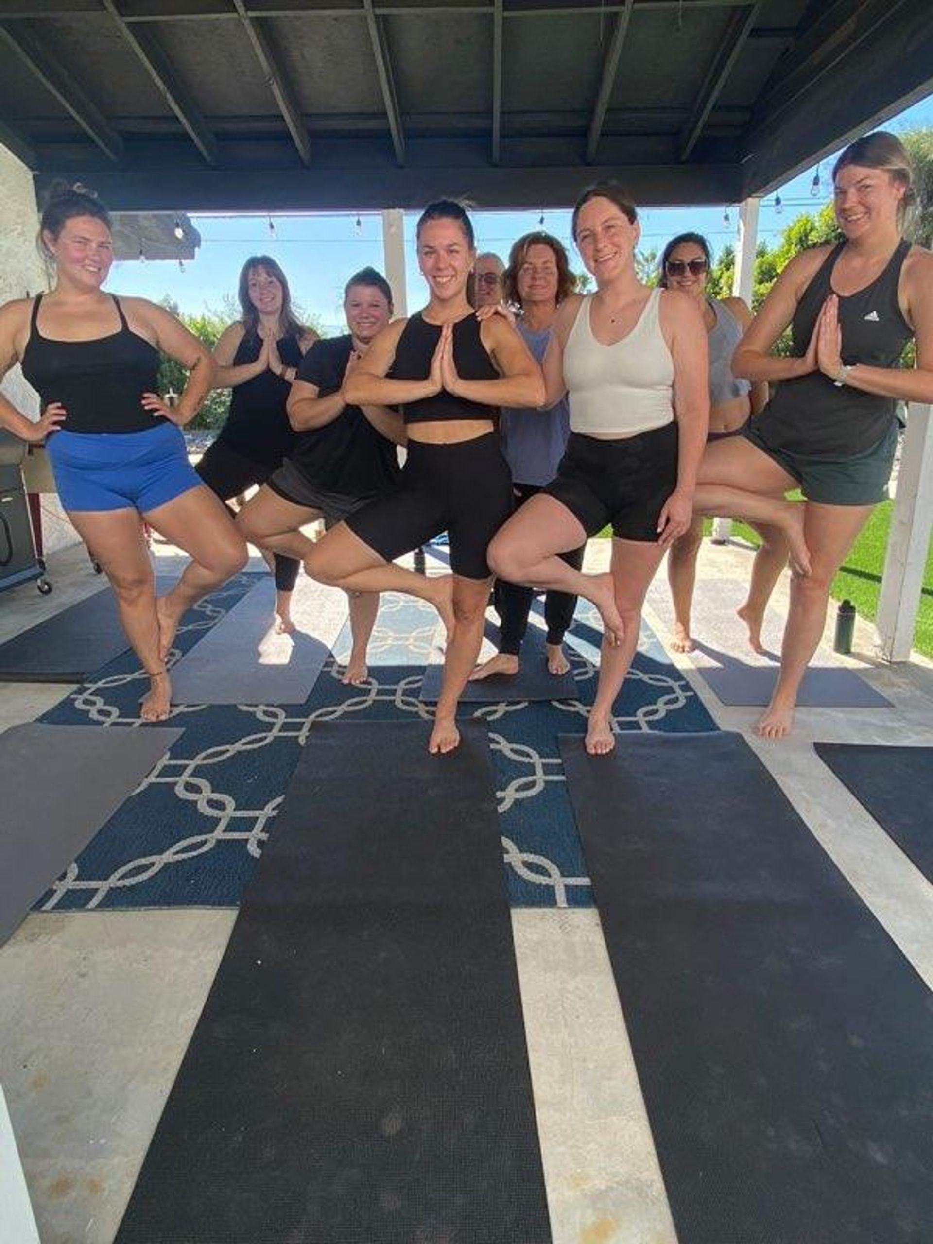 Outdoor patio yoga class: a smiling group of women balancing in tree pose on yoga mats under a shaded pergola in a sunny backyard.