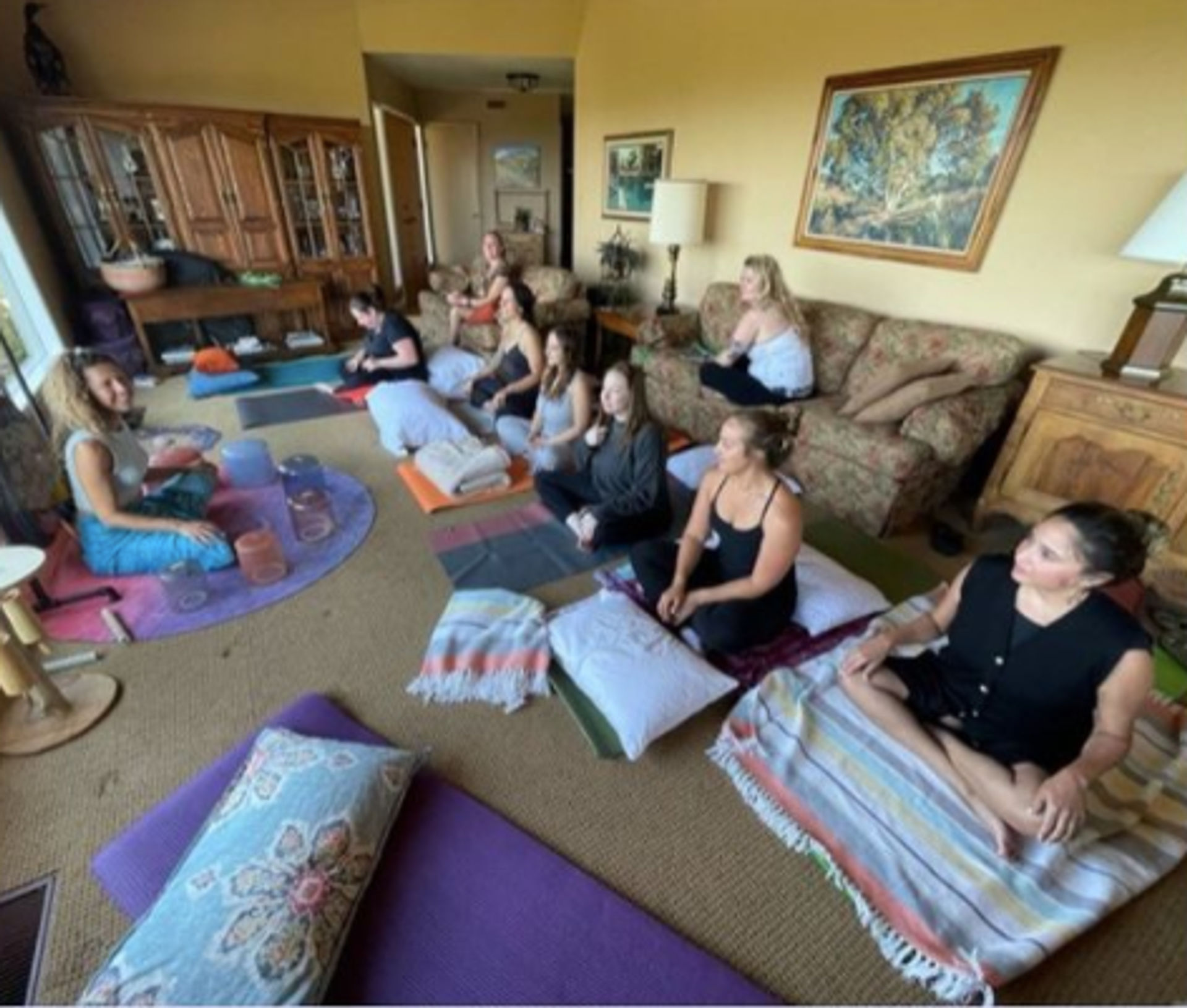 Cozy indoor group meditation and yoga workshop in a living-room setting with women seated on cushions and mats, blankets, warm yellow walls, wooden furniture and framed art.
