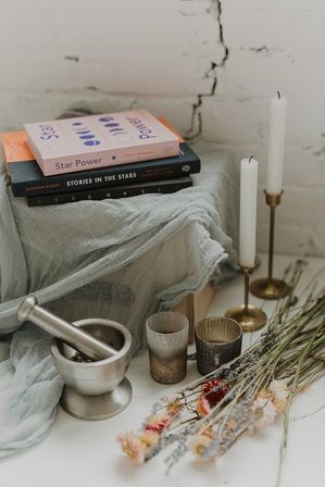 Cozy boho still-life: stack of star-themed books on soft gauzy fabric, metal mortar and pestle, two brass candlesticks with tall white candles, rustic votive cups and a bundle of dried wildflowers against a cracked white brick wall.