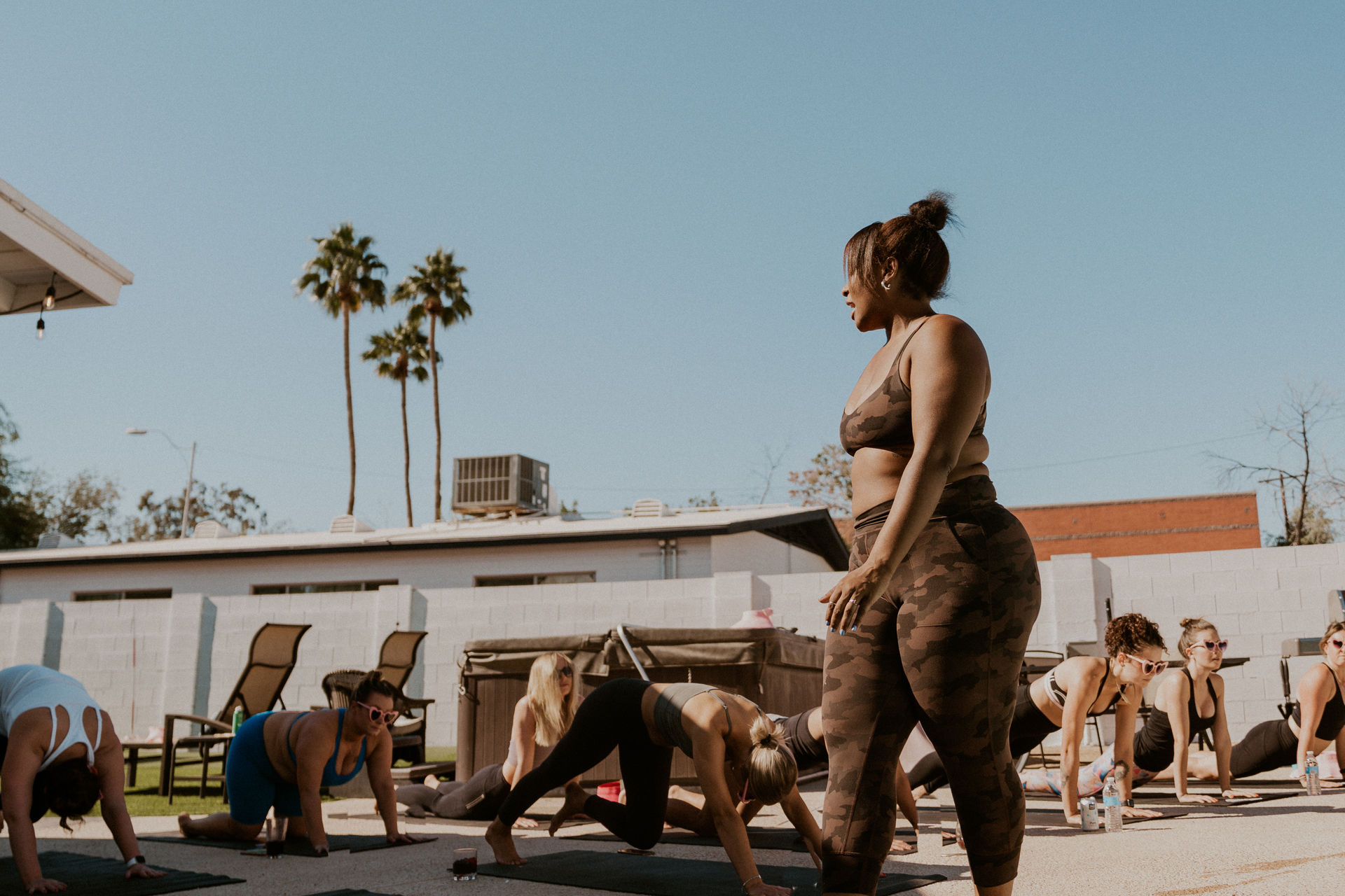 Sunlit outdoor yoga class on a patio in Southern California — instructor in camo activewear leads participants on mats beneath tall palm trees and a clear blue sky.