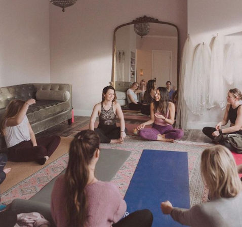 Women’s group yoga class in a cozy indoor studio — participants seated on colorful mats in a circle, smiling and chatting under soft natural light with a vintage mirror and sofa in the background.