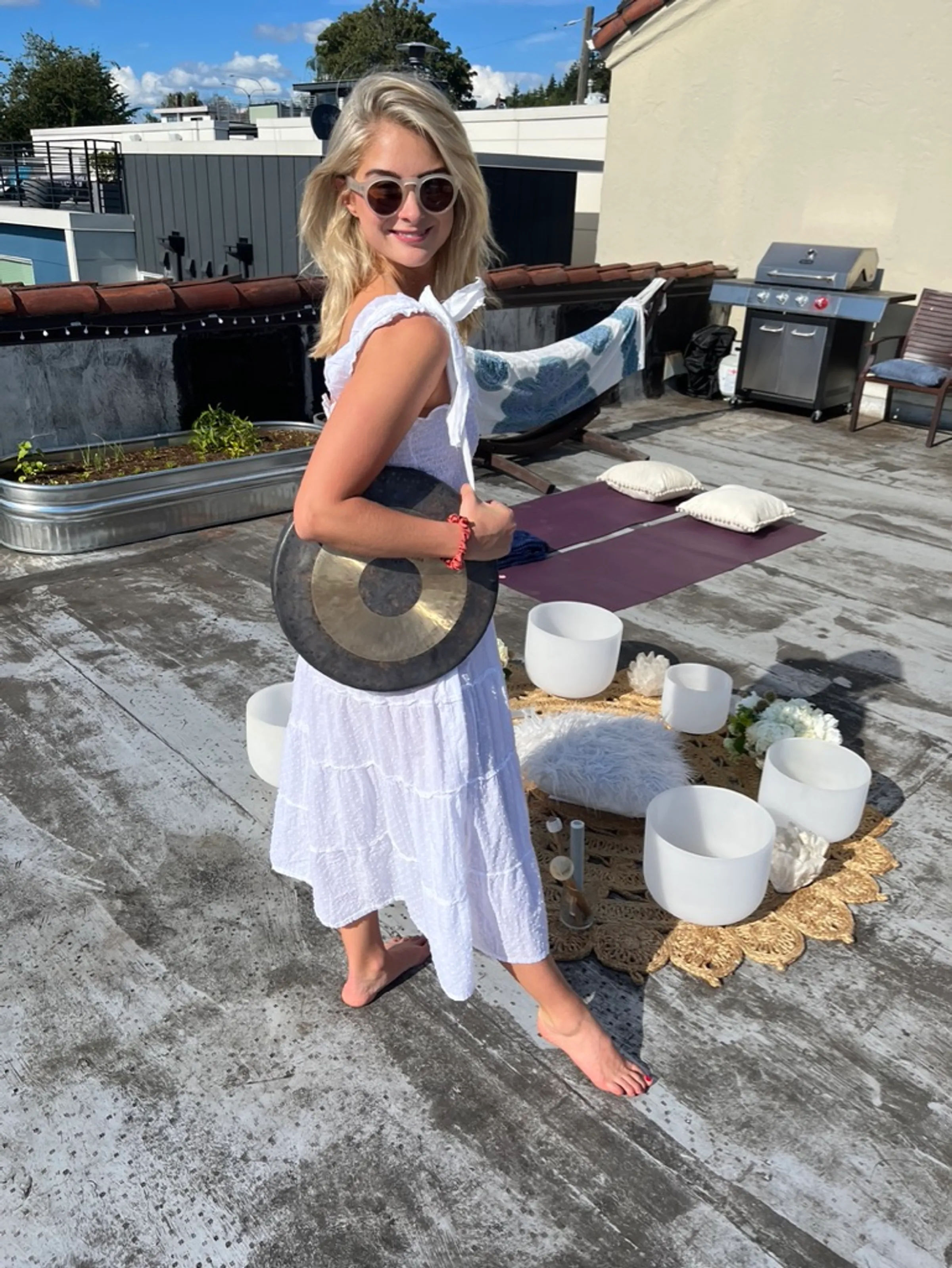 Woman in a white sundress and sunglasses holding a gong on a sunny urban rooftop next to crystal singing bowls, yoga mats and a hammock — rooftop sound bath setup.
