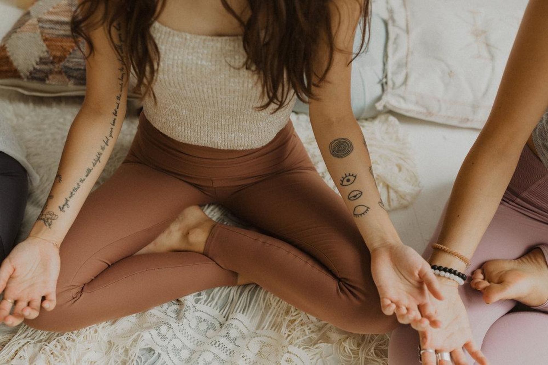 Three people seated cross-legged in a home yoga meditation session, hands resting on knees, forearm tattoos and beaded bracelets visible, wearing neutral leggings and cozy boho textiles