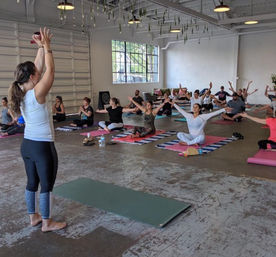 Group yoga class in an urban warehouse-style studio, instructor leading seated participants on colorful mats with arms outstretched beneath hanging plants and a large window