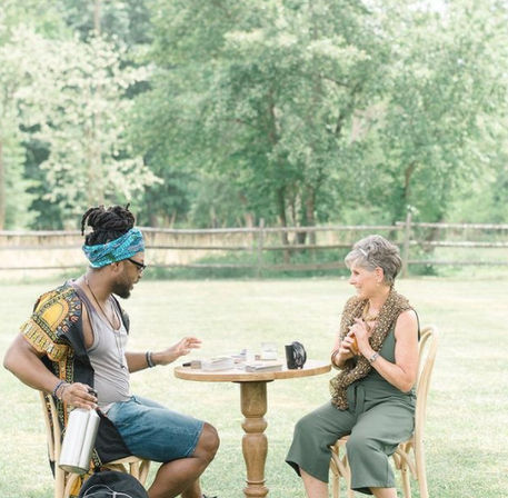 Smiling man and woman sit at a small round table on a sunny grassy lawn, sharing coffee and cards with trees and a rustic wooden fence in the background.