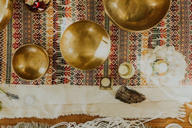 Top-down view of a boho altar: brass Tibetan singing bowls, candles in glass holders, a dark crystal and dried flowers arranged on a colorful geometric woven textile with fringe