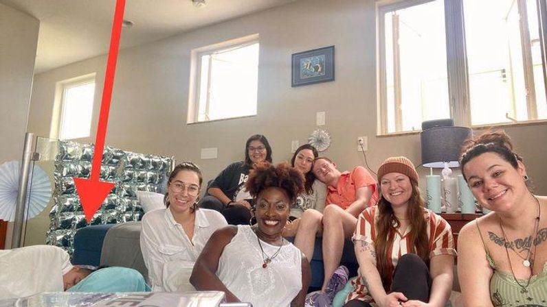 Casual house gathering of friends smiling and lounging on a couch and floor in a sunlit living room, red arrow overlay pointing to someone lying down.