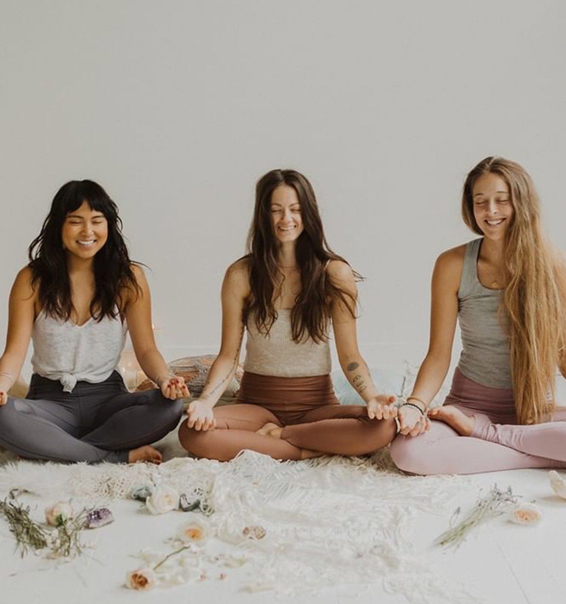 Three women seated cross-legged in a light-filled wellness studio, holding hands and smiling with eyes closed during a calm group meditation on a fluffy rug with scattered flowers.