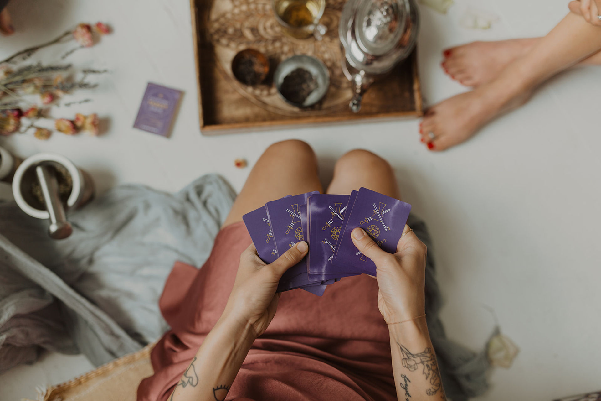 Hands shuffling purple tarot cards over crossed legs in a cozy at-home tarot reading scene with tea, dried flowers and boho decor