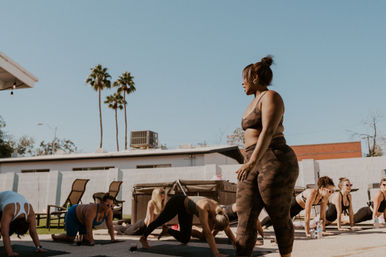 Outdoor yoga class in a sunny courtyard with palm trees — instructor in camo activewear leading group fitness stretches on mats