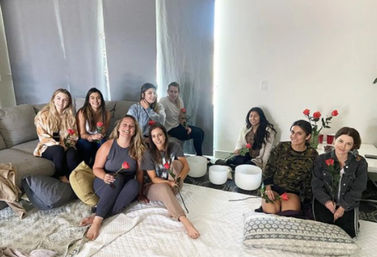 Nine women gathered in a cozy living room for a sound-bath meditation, seated on a sofa and floor cushions holding red roses around white crystal singing bowls.