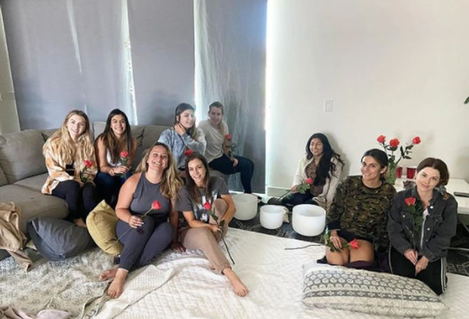 Nine women gathered in a cozy living room for a sound-bath meditation, seated on a sofa and floor cushions holding red roses around white crystal singing bowls.