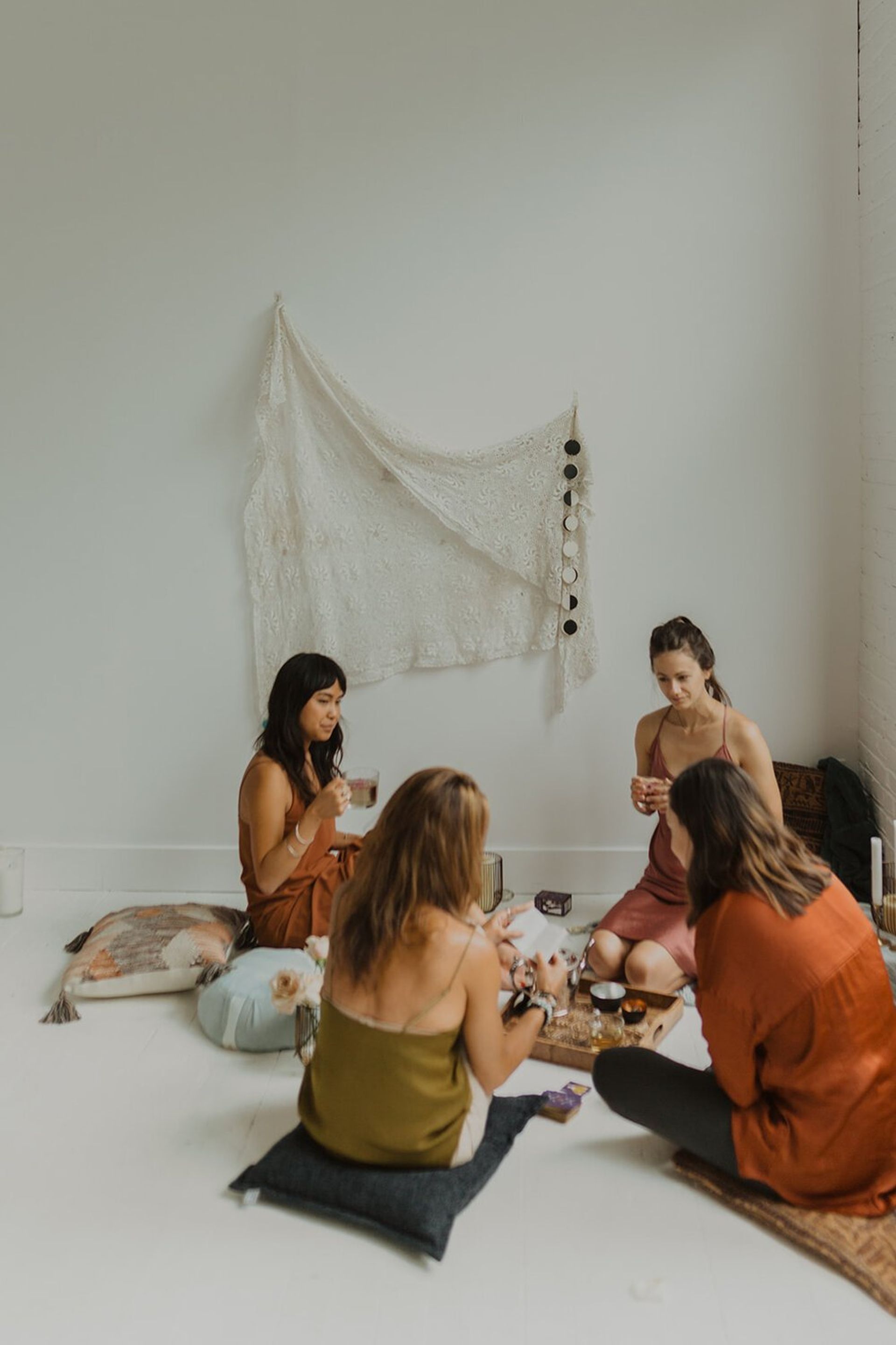 Four women sitting on floor cushions in a sunlit minimalist studio, sharing tea and snacks around a wooden tray with boho textiles and a lace wall hanging — cozy, relaxed indoor gathering.