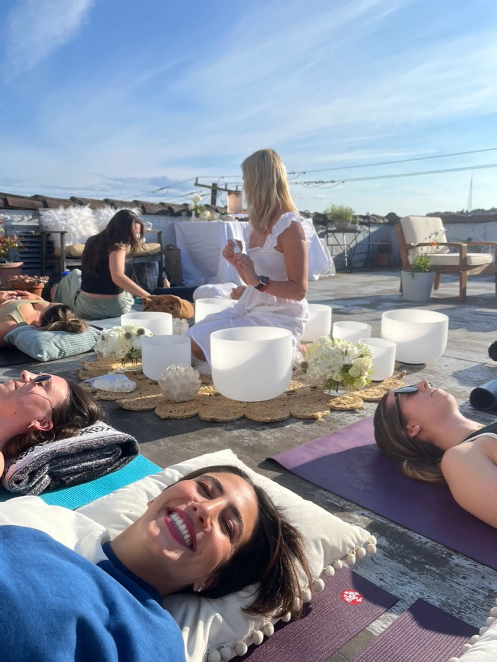 Sunny urban rooftop sound bath with people resting on yoga mats while a woman in white plays white crystal singing bowls amid cushions, flowers and a bright blue sky.