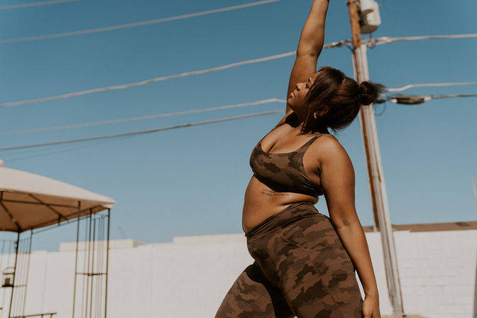 Confident woman in camo workout set stretching one arm up during an outdoor urban yoga session under a clear blue sky with power lines and a white wall backdrop