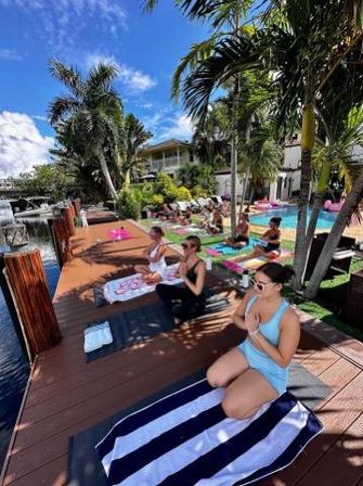 Outdoor yoga class on a sunny tropical waterfront deck by a pool and canal, participants on towels and mats surrounded by palm trees and blue sky