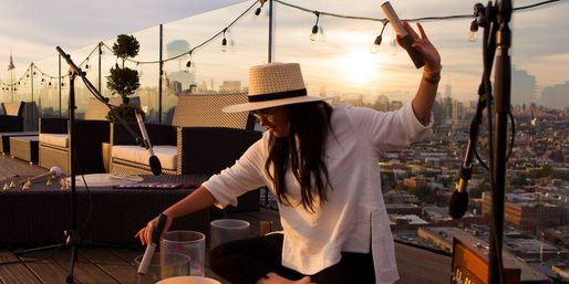Rooftop musician in a straw hat playing crystal singing bowls at sunset, surrounded by microphones, string lights and wicker seating with a city skyline backdrop.