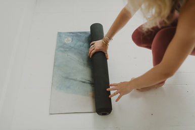 Person rolling up a blue-gray yoga mat on a bright white wooden floor, prepping for an indoor yoga session.