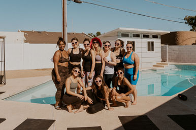 Smiling women in activewear and heart-shaped sunglasses pose together on a sunny suburban backyard pool deck for a playful pool-party and fitness gathering.