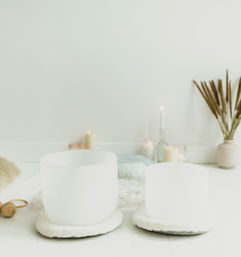 Minimalist meditation space with two white crystal singing bowls on plush cushions, lit candles and dried reeds for a calming sound‑healing setup.