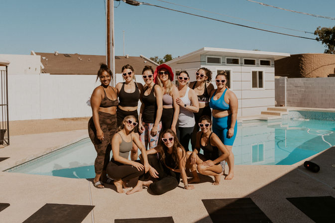 Ten women in colorful activewear and heart-shaped sunglasses pose poolside at a sunny suburban backyard pool and modern pool house — outdoor fitness meetup.