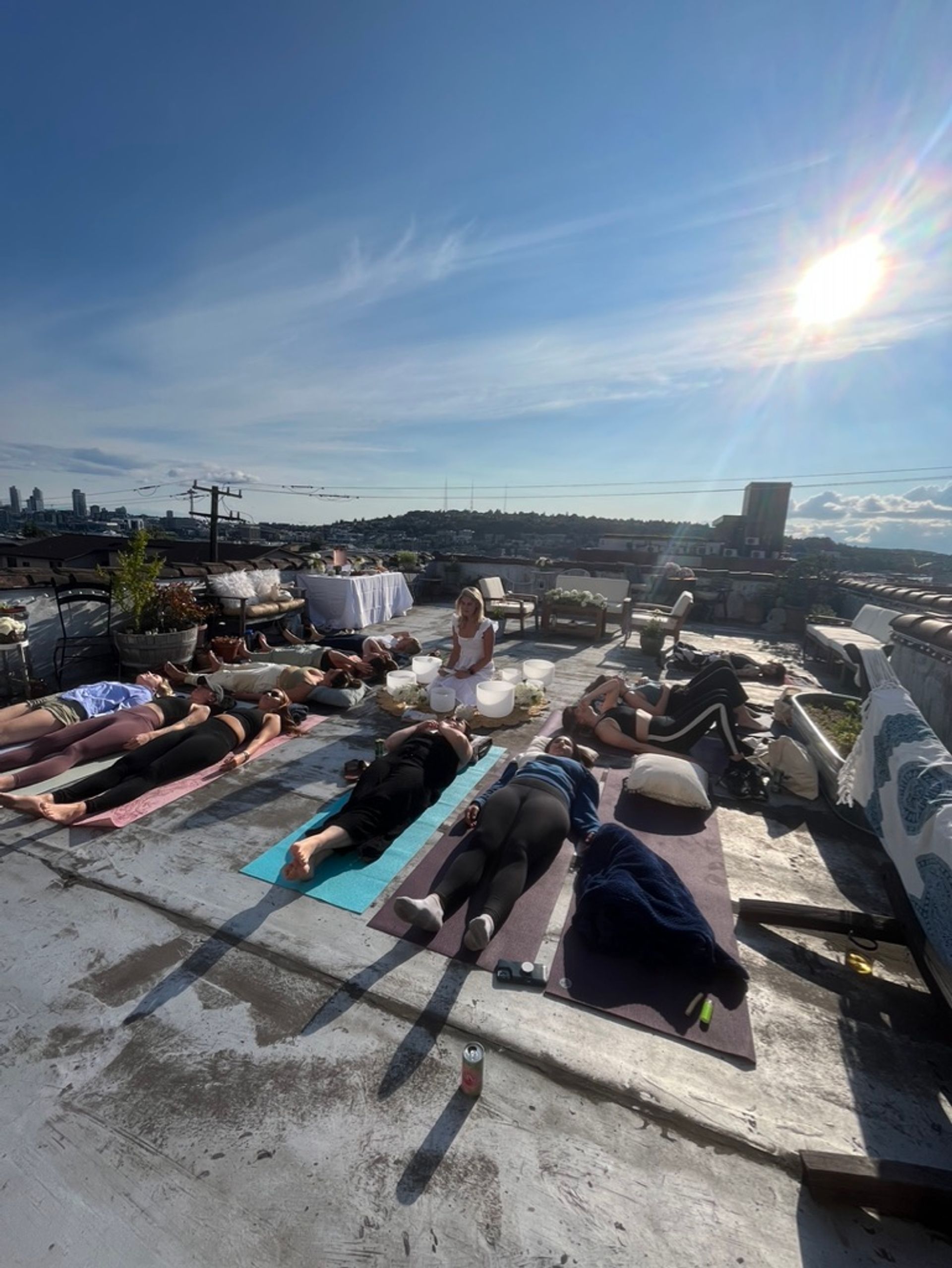 Group rooftop sound-bath yoga on an urban rooftop — people resting on mats in Savasana with crystal singing bowls and a bright sunlit skyline.