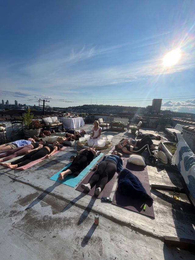Group rooftop sound-bath yoga on an urban rooftop — people resting on mats in Savasana with crystal singing bowls and a bright sunlit skyline.