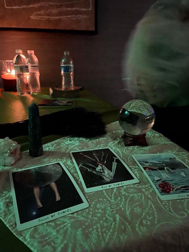 Dimly lit indoor tarot reading setup on a table with three illustrated tarot cards (Seven of Swords, Ten of Swords, Five of Cups), a clear crystal ball on a wooden stand, raw crystals and a tall dark obelisk, candlelight and water bottles in the background.