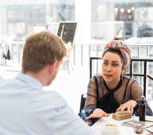 Tarot card reader with colorful headscarf doing a reading for a customer at a bright urban cafe table, tarot cards and crystals visible.