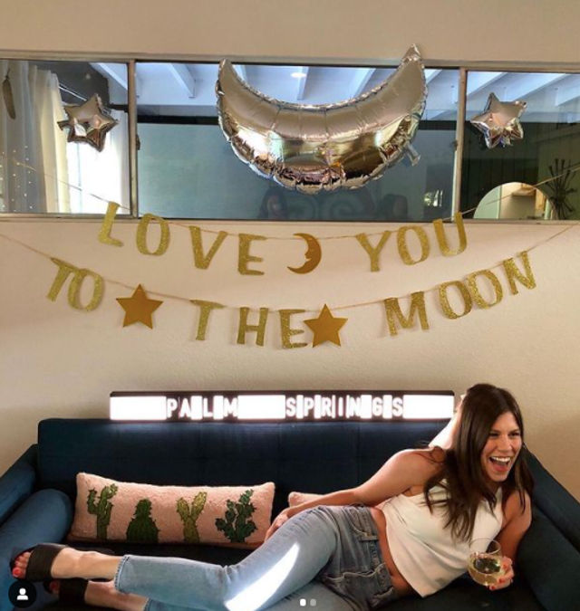 Laughing woman lounging on a blue sofa with a drink at an indoor celebration under a gold “Love You to the Moon” garland, silver crescent and star balloons, cactus-print pillows and an illuminated lightbox.