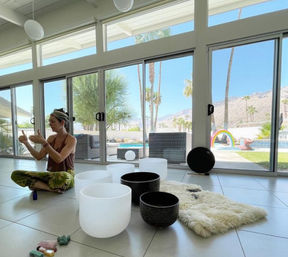 Woman seated cross-legged among white and black singing bowls and crystals in a bright sunroom with floor-to-ceiling windows overlooking palm trees, a pool with a rainbow float and desert mountains.
