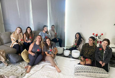 Group of nine women in a cozy living room sitting on a couch and floor with cushions and rugs, each holding a red rose beside white crystal singing bowls for a relaxed wellness/meditation gathering.