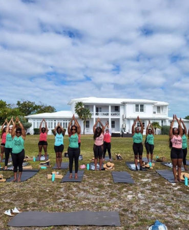Outdoor group yoga class on mats across a grassy lawn in front of a large white coastal house with palm trees, water bottles and hats visible under a bright, partly cloudy sky.