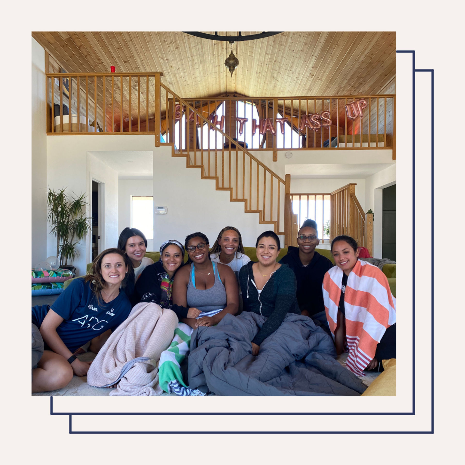 Group of eight friends smiling and wrapped in blankets on a living room floor of a bright vacation-style home with a wood-paneled vaulted ceiling, open staircase and loft railing decorated with a festive balloon banner.