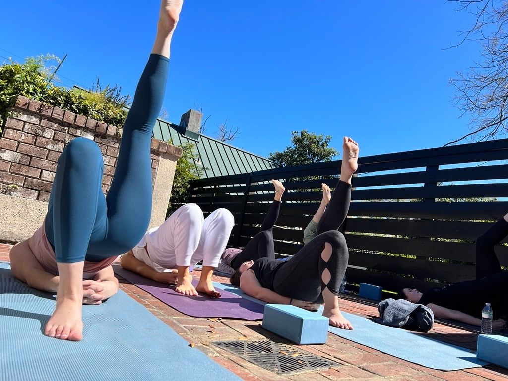 Sunny outdoor yoga class on a brick patio with participants on mats doing bridge and single-leg lifts, legs raised toward a bright blue sky.