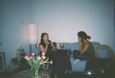 Two people chatting on a blue couch in a cozy living room at night, soft floor lamp and wine glasses on a small table, pink tulips in a vase on the coffee table in the foreground.
