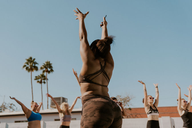 Outdoor group fitness class in sunny Southern California — instructor in camo leggings and strappy sports bra leads participants with arms raised beneath palm trees and a clear blue sky.